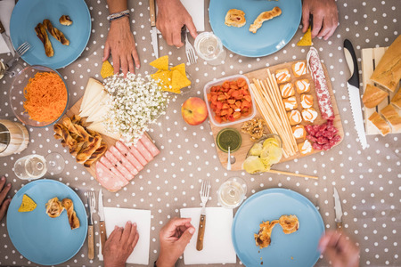 highview of a table during a lunch for 4 senior men and women. bright image with food like mortadella, carrots, slami, bread and more.many hands on the table waiting to eatの写真素材