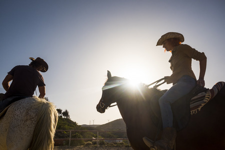 couple of modern cowboys man and woman ride two horses outdoor with sunflare and backlight. mountains and wind mill in background. nice young on vacationの写真素材
