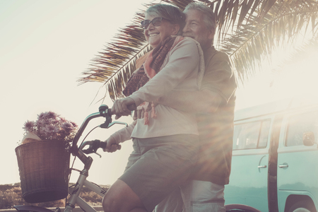 beautiful aged couple of senior in vacation go both on an old bike and enjoy the summer sunlight. tropical place and vintage van in background. retro colors and style for happiness conceptの写真素材