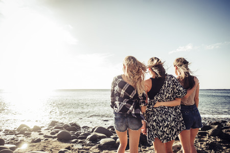 Group of three young nice girls embrace themselves looking at the ocean sunset in vacation. Team moment full of emotions and memories. Sunlight in a blue sky.の写真素材