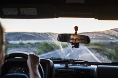 nice caucasian adult lady drive the car with dirty glass. long way road traveling to the sunset in front. outdoor nature and mountains. looking the back mirror to check the trafficの写真素材