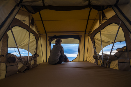 lonely independent strong middle age woman feeling the nature outdoor in a roof tent on the car. travel and lifestyle wanderlust concept for beautiful caucasian lady sitting in front of the oceanの写真素材