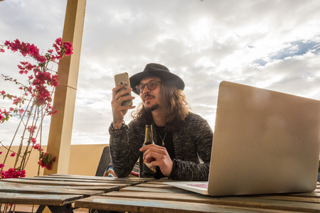 young happy man work with smartphone and laptop on the table and drinking beer. alternative office station and technology lifestyle for caucasian model sitting and enjoying internetの写真素材