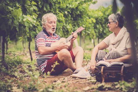 couple of alternative aged older traveler stay sitting down in a vineyard with the luggage and playing an ukulele acoustic guitar and reading a paper book.の写真素材