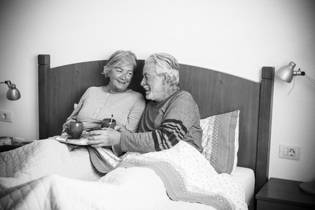caucasian aged couple doing breakfast at home in the bed. nice natural scene at home for togheterness life concept. love and carefree people married. laughing together starting the day in the morningの写真素材