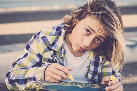 beautiful young boy teenager with long blonde hair trendy shirt write enjoy on a recycled piece of wood. freedom and alternative lifestyle for attractive guy. looking to the camera portraitの写真素材