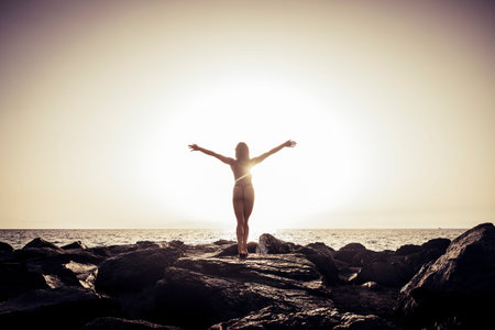 Rear view of a beautiful, brunette young girl with raised hands, looking at ocean. Freedom concept, holiday, beach, clear sky background., light and shadow landscape scenery placeの写真素材