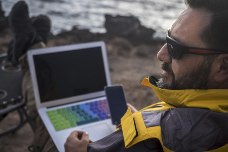 modern digital nomad traveler bearded man work with mobile phone and laptop outdoor in front of the ocean. alternative office concept for free male and adventure lifestyle, sunglasses and feel of nature for travel journeyの写真素材
