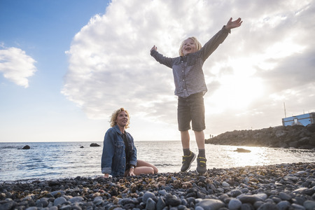 Happiness concept with blonde young mother sitting at the beach while little child son jumping full of joy near her.の写真素材