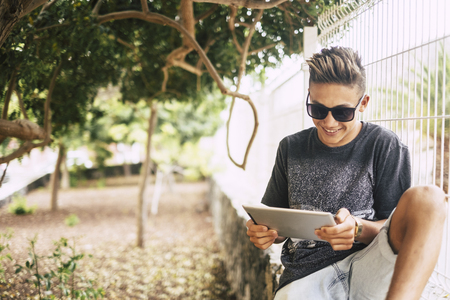 young millennial teenager use tablet with internet and wifi outdoor at the park to connect and see his friends at home.の写真素材