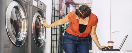 caucasian young lady use notebook computer at the laundry mat automatic 24 h service shop while wait for her new washed and cleaned clothes. internet wifi business activity for millennial casual peopleの写真素材