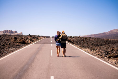 nice young couple viewed from rear walking together hugging on a long way road in the middle of the lava desert on an asphalt road. clear sky in background and arid landscape around them. love and friendship conceptの写真素材