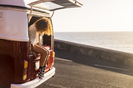 wanderlust and vanilife concept with young caucasian woman traveler sitting out of a vintage retro legenday van ready to travel the world. sunset and ocean in front of her parked just close to the sea. freedom totallyの写真素材
