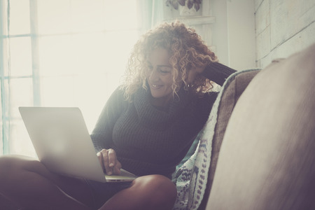 Curly woman with gray sweater sitting on brown sofa and blue blankets relaxes looking at the computer on an autumn day. Technology conceptの写真素材