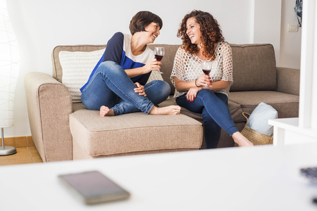 Couple of women friends sit down on the couch together enjoying the afternoon and the friendship with a cup of red wine laughing and smiling telling stories. Life home real daily.の写真素材