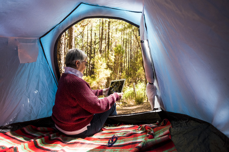 Mature adult retired woman sitting inside a tent in free wild camping alone in the forest using a technology internet connected tablet to organize the travel and check social media for digital nomad workの写真素材