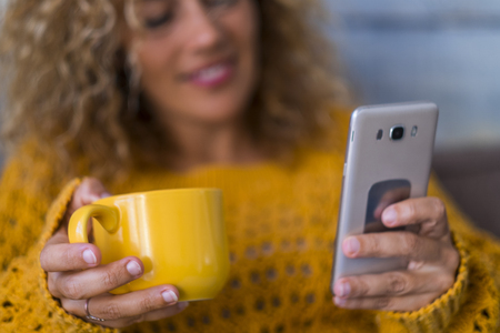 Autumn and technology mobile phone cconcept - middle age modern woman drink tea or coffee at home while chek the cellular for internete messages od news from social media - focus on mugの写真素材