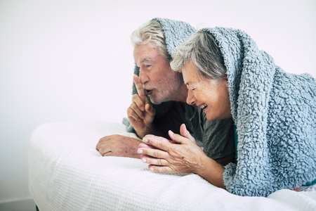 Happy adult couple have fun laughing together at home in the nedroom under the sheet in the wake up morning time - together forever with happiness concept for caucasian people in loveの写真素材