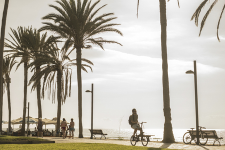 young man ride on a bike during summer warm day of vacation on the coastline alone with people in bikini around walking enjoying the holiday time. real life scene for happy people in outdoor leisures activitiesの写真素材