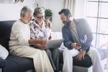 Caucasian aged family at home matures and middle age people together using technology devices like laptop and mobile phone enjoying internet web results - everyday lifestyle for modern men and womenの写真素材