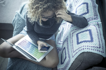 Curly woman and glasses spends time chatting with the phone sitting on the sofa in the comfort of her home. Technology and laptop device. Alternative office and work station for independent beautiful middle age womanの写真素材