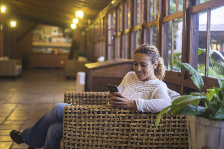 woman sitting on wicker chair in wooden house with large picture window smiling looking on cellphone. Tehcnlogy leisure activity for cheerful people indoorの写真素材