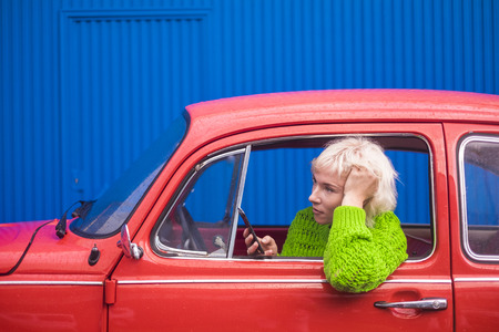 Blonde beautiful woman 30 years old inside her red vintage car using technology modern mobile phone in a rainy day. Colors and travel lifestyle for millennial lady conceptの写真素材