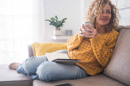 Beautiful 40 years old caucasian lady sit down on the sofa drinking tea and reading a book for afternoon indoor leisure activity at home - after work lifestyle for people conceptの写真素材
