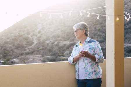 White hair aged beautiful woman drink a cuo of white wine outdoor in the terrace looking down and enjoying the sunset at home on the rooftop with mountain viewの写真素材