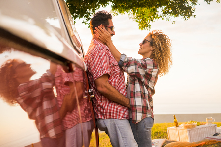 Couple in love middle age caucasian beautiful people hug during a picnic and vacation travel or outdoor leisure activity together with old red vingtage van to discover the world - hiupster style for traveler - happiness conceptの写真素材