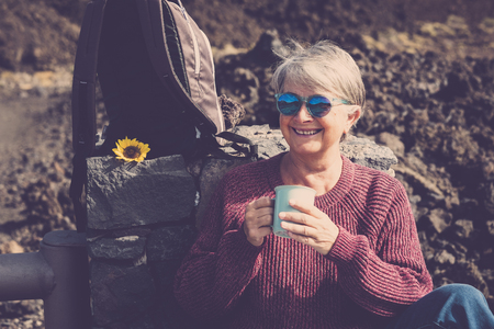 Cheerful happy caucasian aged mature woman enjoy a hot beverage like tea or coffee resting after a trekking day of outdoor leisure activity at the mountain - alternative vacation lifestyleの写真素材