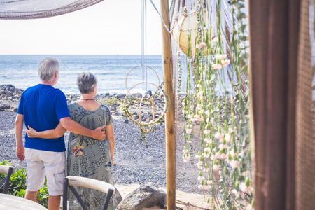 Retired couple in relationship looking at the sea during summer spring vacation together - happy leisure outdoor activity for married aged people with ocean in background - tourists concept at the beachの写真素材