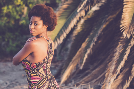 Fashion portrait of black traditional coloured dress african model pose with tropical palm in background. Ethnic beauty with afro hair style loooking to you outdoor in a sunny dayの写真素材