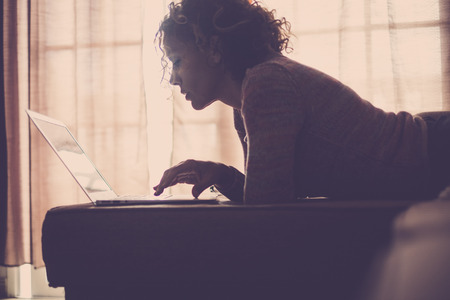 woman with window backlightand jeans lying on sofa working with laptop computer in complete home relaxation. Freedom technology concept and alternative office for digital nomad and modern freelancersの写真素材