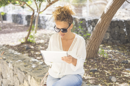 Beautiful middle age woman working outside in the park with a modern tablet using internet and touchscreen display device. Outdoor office in a laptop concept for free people digital nomadの写真素材