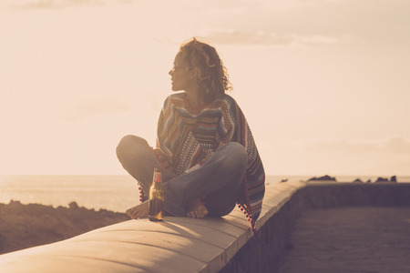 Attractive strong independent woman sit down on a wall barefoot and hippy style looking at the nature around and enjoying a golden outdoor leisure lifestyle sunset near ocean - coloured lifestyleの写真素材