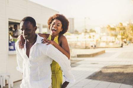 Happy cheerful attractive black skin african couple enjoy the outdoor leisure activity in the city. The man carry the laughing girl on his back. Beautiful millennial people love and play togetherの写真素材