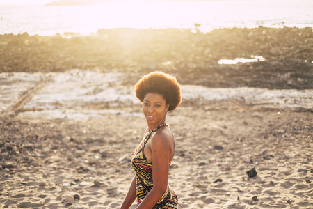 Beautiful african hair skin race lady on the beach with sunset and backlight in background enjoying outdoor leisure activity. Traditional dress afro style and colors. Beautiful black modelの写真素材