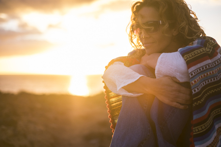 romantic portrait for freedom and peace concept with beautiful middle age caucasian lady stay sit down with mexican poncho and golden light from sunset in background - enjoying lifeの写真素材