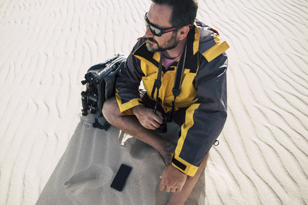 explorer  man sitting and resting in a desert adventure. sand everywhere. photographer looking for beautiful places destination. moern phone on the ground and backpack ready to cross the dunes and enjoyの写真素材