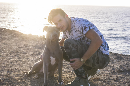 Portrait of young beautiful attractive young man and his best friend amstal dog sit down with him - ocean and scenic sunlight in background - pet therapy and love for animals conceptの写真素材