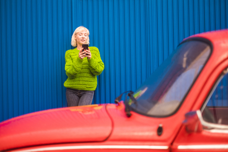 Fashion coloured concept image with blonde caucasian girl standing near a red vintage old car with blue steel wall in backgorund. Urban and colors with people travel technology conceptの写真素材