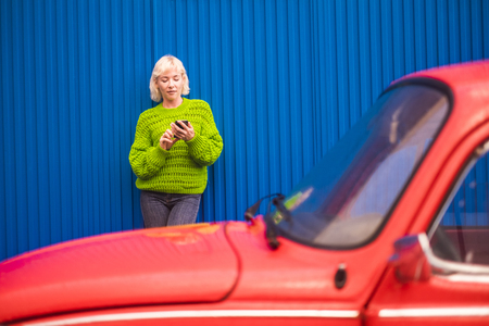 Colors and alternative happy lifestyle concept image with blonde attractive woman thirty years old using mobile phone internet technology outside the red car and with blue wall on the backgroundの写真素材