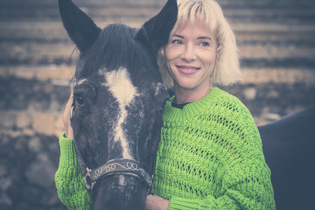Beautiful european young blonde lady with green sweater hug with love a black horse for pet therapy - animals best friends forever together with nice people outdoorの写真素材