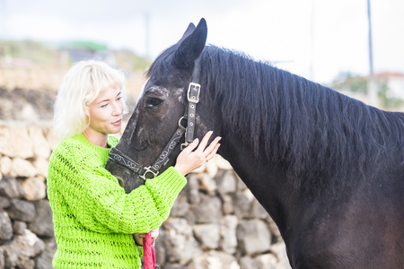 Horse therapy love concept with female blonde young caucasian lady and black lovely domestic horse hugging and stay in friendship outdoor during the day - enjoy the alternative leisure activity riding animalの写真素材