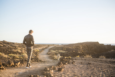 Lonely man and best friend dog walking together on a path in outdoor desert leisure activity with ocean in background at the end - friendship and travel concept for people with animalsの写真素材