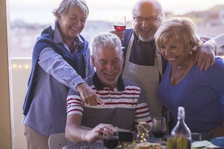 Adults senior caucasian friends have fun taking and looking piucrures on smart phone during friendship dinner together outdoor - laughing and smiling sharingfun with technology deviceの写真素材