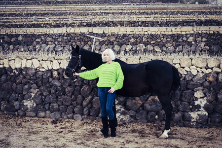 Fashion concept with cute blonde model posing in the countryside with a black horse and wearing a green jacket. colors and contrast people and animalsの写真素材