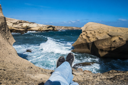 Travel and discover scenic places concpet for legs lay down and relax on a cliff with blue big and dangerous storm ocean waves - colors and coast activity in vacationの写真素材