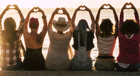 Love and meditation lifestyle concept - group of women looking at the sunset on the ocean and make the hearth with hands - romantic golden light and valentine's day concept - friendshp forever